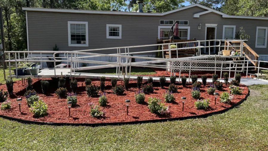 A beige mobile home with a long metal wheelchair ramp leading to the front door, surrounded by a red mulch flowerbed.